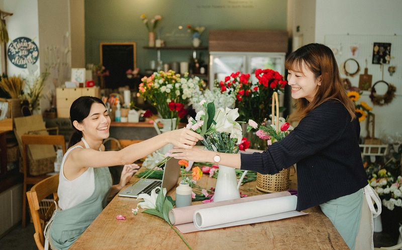 Two smiling women in a flower shop exchanging flowers, representing Seattle businesses fostering a culture of giving and authentic community support through meaningful partnerships and local initiatives.